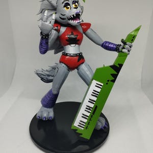 May include: A figurine of a wolf-like character with gray fur, a red outfit, and a green keytar. The figure is on a black base, with a green mohawk and purple accents. The character is making a hand gesture.