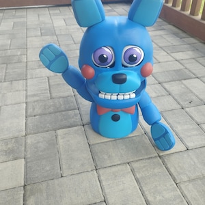 May include: A blue bunny figure with large ears, purple eyes, and a red bow tie. The figure has a friendly expression with a wide smile and is waving one hand. The figure is made of a smooth material and is standing on a gray brick surface.