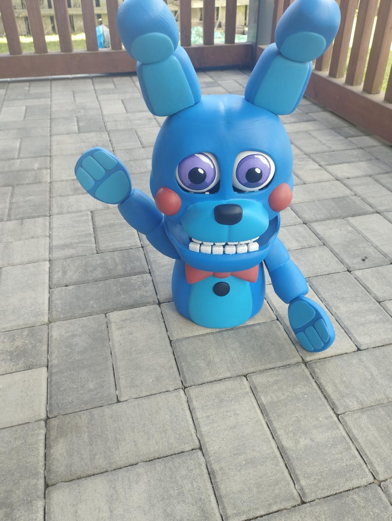 May include: A blue bunny figure with large ears, purple eyes, and a red bow tie. The figure has a friendly expression with a wide smile and is waving one hand. The figure is made of a smooth material and is standing on a gray brick surface.