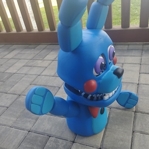 May include: A blue bunny figurine with large ears, a red bow tie, and a black nose. The figurine has a wide grin with visible teeth and outstretched arms. The eyes are purple with white highlights. The toy is on a gray brick surface.
