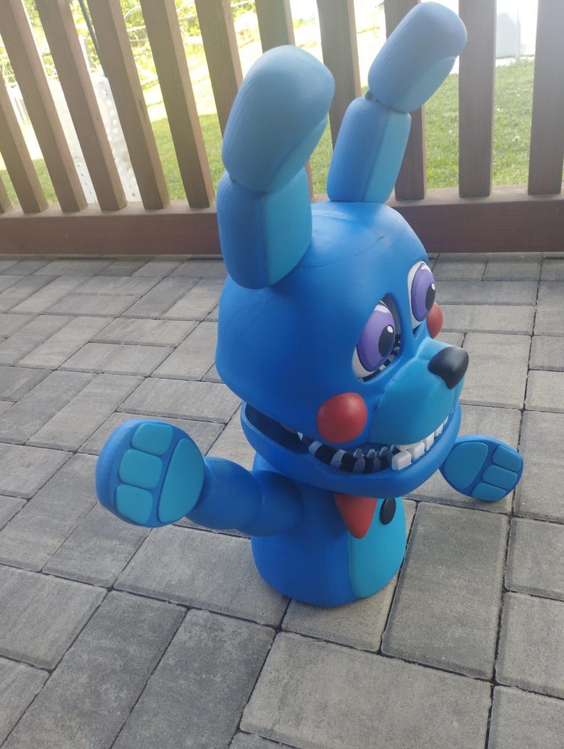 May include: A blue bunny figurine with large ears, a red bow tie, and a black nose. The figurine has a wide grin with visible teeth and outstretched arms. The eyes are purple with white highlights. The toy is on a gray brick surface.