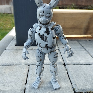 May include: A gray, weathered, and damaged animatronic figure with bunny ears, a wide grin, and a missing eye. The figure is standing on a gray concrete surface.