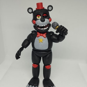 May include: A black and grey bear figurine with a red top hat and bow tie, holding a microphone. The bear has a grey chest with a gold star, red accents on the knees, and grey feet. The figurine is a collectible toy.