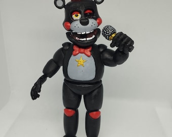 Puede incluir: Figura de oso negro y gris con sombrero de copa y pajarita rojos, sosteniendo un micrófono. El oso tiene un pecho gris con una estrella dorada, detalles rojos en las rodillas y pies grises. Juguete coleccionable.