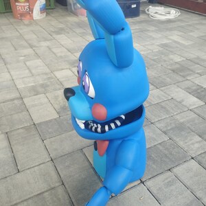 May include: A large, blue bunny figure with articulated limbs, large ears, and a red bow tie. The bunny has a black nose, pink cheeks, and purple eyes. The mouth is open, revealing white teeth. The figure is likely a character from a video game or cartoon.