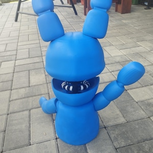 May include: A large, bright blue, cartoon-like figure of a rabbit with large ears and outstretched arms. The rabbit has a round head and body, with a visible mouth revealing white and black striped teeth. The figure is standing on a gray brick surface.