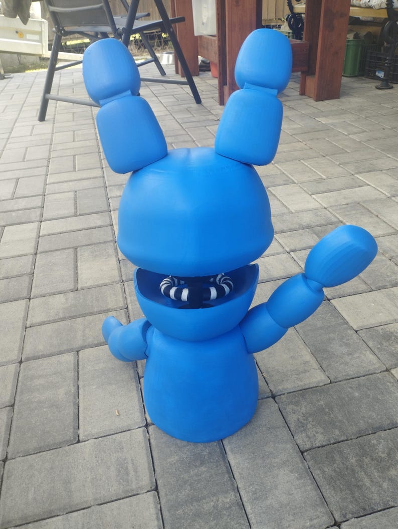 May include: A large, bright blue, cartoon-like figure of a rabbit with large ears and outstretched arms. The rabbit has a round head and body, with a visible mouth revealing white and black striped teeth. The figure is standing on a gray brick surface.