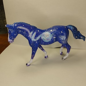 May include: A blue and white painted horse figurine with a galaxy design. The horse has a white mane and tail and is standing on a white surface.
