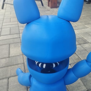 May include: A bright blue, three-dimensional figure of a rabbit with large ears and outstretched arms. The rabbit has a wide open mouth with a black and white striped interior. The figure is made of a solid material and is standing on a brick surface.