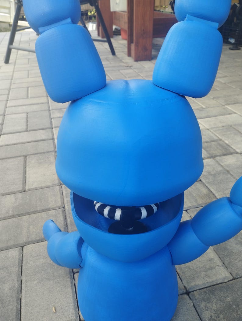 May include: A bright blue, three-dimensional figure of a rabbit with large ears and outstretched arms. The rabbit has a wide open mouth with a black and white striped interior. The figure is made of a solid material and is standing on a brick surface.
