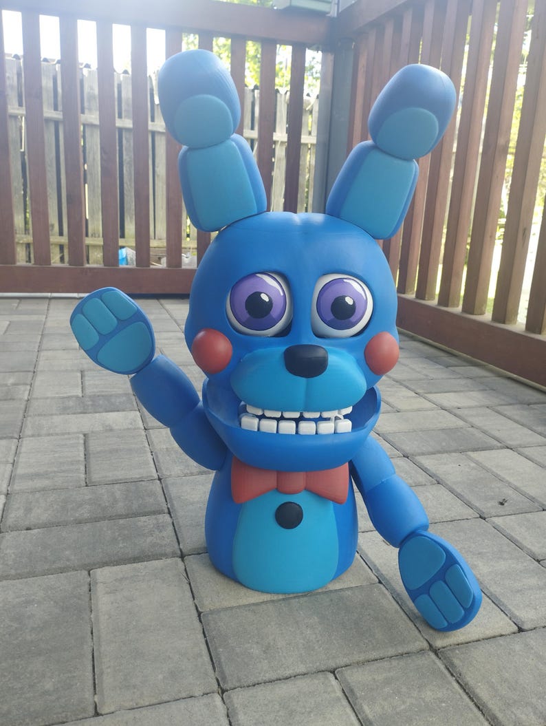 May include: A blue bunny figure with large ears, a red bow tie, and a friendly expression. The figure has large purple eyes, red cheeks, and a wide, toothy grin. The figure is standing on a gray brick surface.