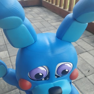 May include: A blue bunny figure with large ears, red cheeks, and purple eyes. The figure has a black nose and a wide, toothy grin. The toy is made of a solid material and is positioned outdoors.