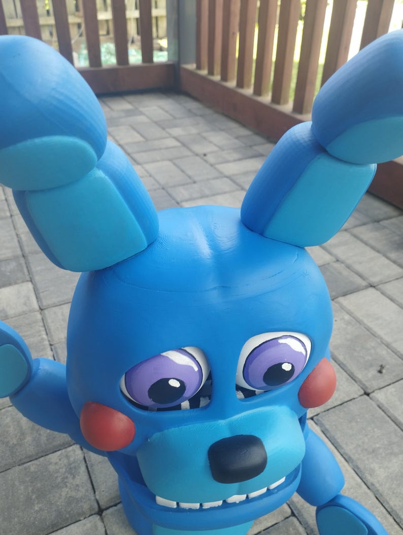 May include: A blue bunny figure with large ears, red cheeks, and purple eyes. The figure has a black nose and a wide, toothy grin. The toy is made of a solid material and is positioned outdoors.