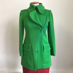 Könnte beinhalten: Eine leuchtend grüne Vintage-Jacke mit einem diagonalen Reißverschluss und zwei großen Taschen. Die Jacke hat einen hohen Kragen und lange Ärmel.
