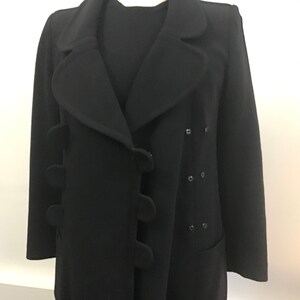 Pu&ograve; includere: Un cappotto di lana nero con chiusura a doppio petto e bottoni arrotondati unici. Il cappotto ha un colletto largo e una singola tasca sul lato destro.