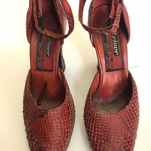 Könnte beinhalten: Ein Paar rote Schlangenlederschuhe mit offener Zehenpartie. Die Schuhe haben einen Riemen um den Knöchel und der Markenname "JUNGE MODE" ist auf der Innenseite des Schuhs aufgedruckt.