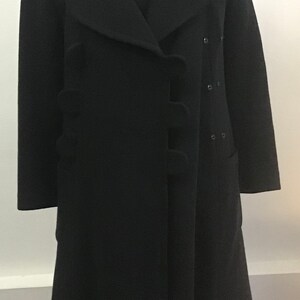 Pu&ograve; includere: Un cappotto di lana nero con un design unico che presenta un davanti con balze e una chiusura con un solo bottone. Il cappotto ha una silhouette lunga e aderente e un orlo leggermente svasato.
