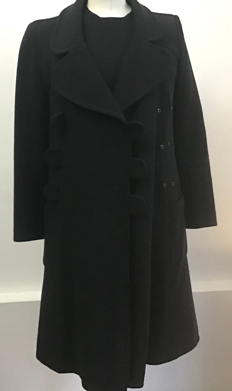 Pu&ograve; includere: Un cappotto di lana nero con un design unico che presenta un davanti con balze e una chiusura con un solo bottone. Il cappotto ha una silhouette lunga e aderente e un orlo leggermente svasato.