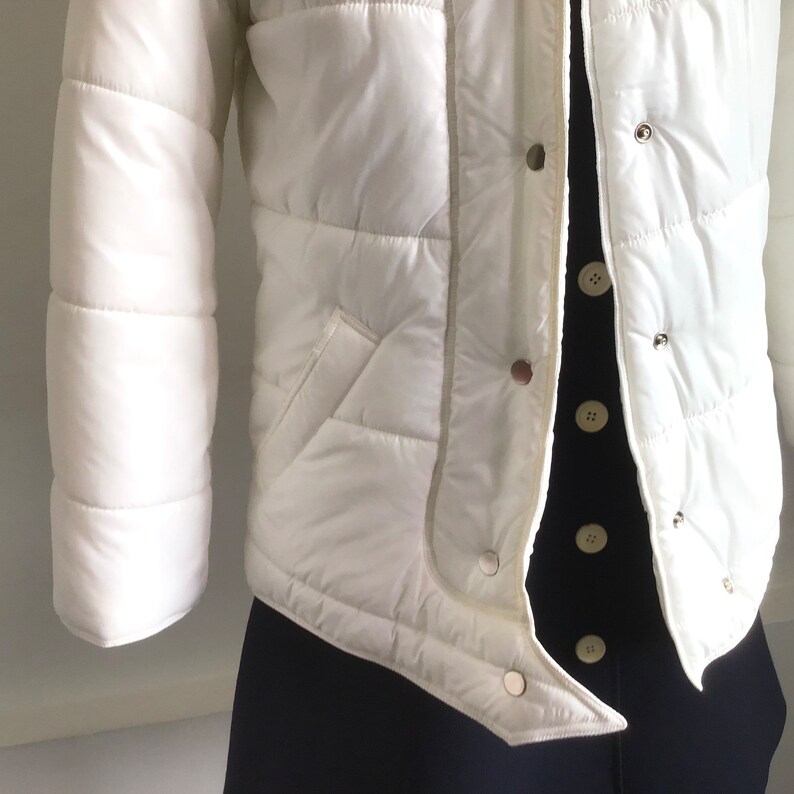 May include: White puffer jacket with a zipper closure and a stand-up collar. The jacket has a quilted design and a white trim around the edges.