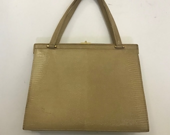 May include: A tan handbag with a textured, reptile-like pattern. The bag has a rectangular shape with a clasp closure and two handles. The hardware is gold-toned. The bag is set against a plain white background.