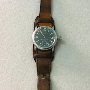 Könnte beinhalten: Eine silberne Armbanduhr mit schwarzem Zifferblatt und braunem Lederband. Das Zifferblatt der Uhr hat weiße Markierungen und den Text "Omega Automatic" und "Anti-Magnetic".