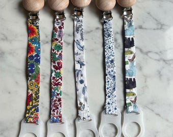 Könnte beinhalten: Fünf Schnullerclips für Babys mit Holzperlen und Stoffbändern mit Mustern. Die Bänder weisen Blumen-, Vogel- und Sterndesigns in verschiedenen Farben auf. Jeder Clip hat eine transparente Kunststoffschlaufe.
