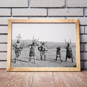 May include: Black and white photograph of five people wearing traditional Native American headdresses and clothing. They are standing in a field and holding weapons.