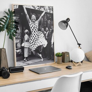 Pode incluir: Fotografia em preto e branco de uma mulher com um vestido de bolinhas pulando no ar com os bra&ccedil;os levantados. A foto est&aacute; emoldurada e pendurada na parede acima de uma mesa com um laptop, uma l&acirc;mpada e uma planta em vaso.
