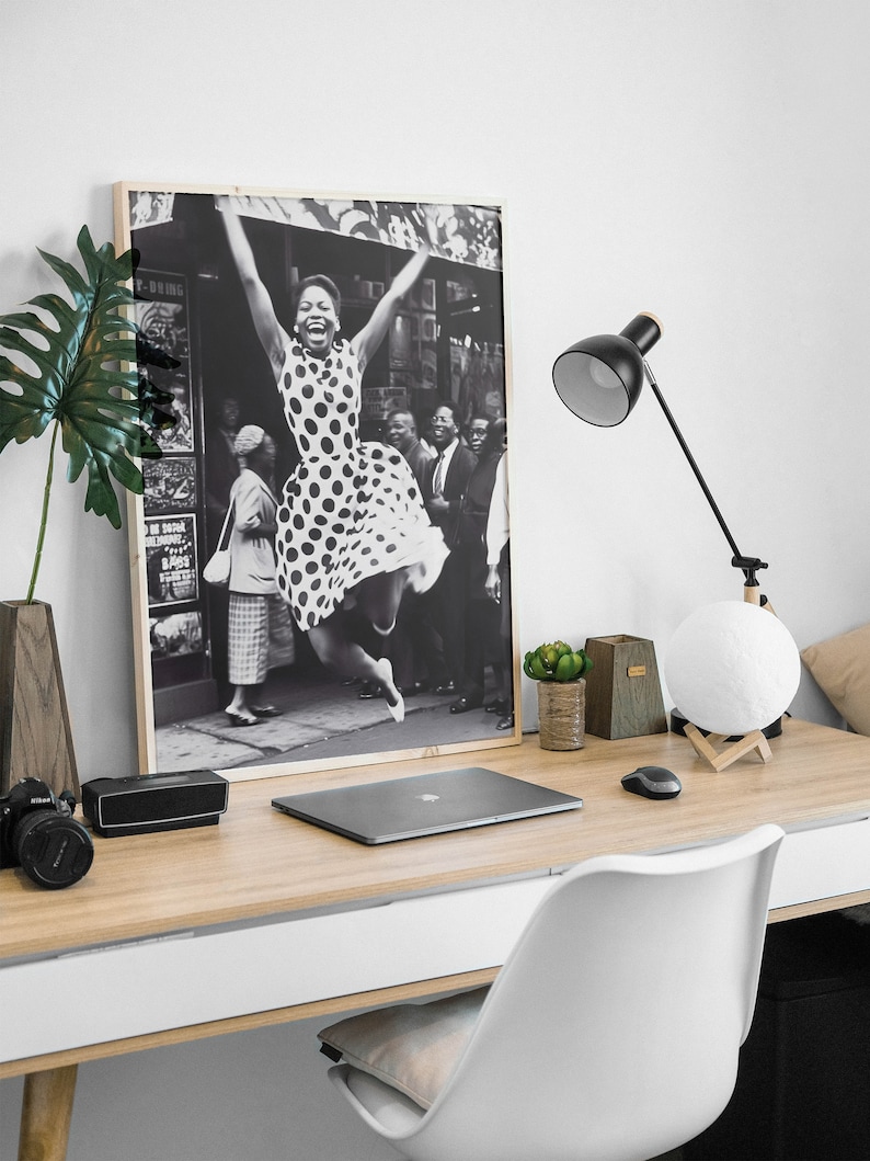 Pode incluir: Fotografia em preto e branco de uma mulher com um vestido de bolinhas pulando no ar com os bra&ccedil;os levantados. A foto est&aacute; emoldurada e pendurada na parede acima de uma mesa com um laptop, uma l&acirc;mpada e uma planta em vaso.