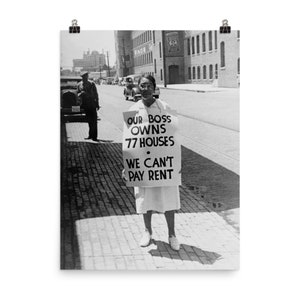 May include: A woman in a white dress holds a sign that reads "Our Boss Owns 77 Houses. We Can't Pay Rent." The sign is in black and white lettering on a white background. The woman is standing on a brick street in front of a building.