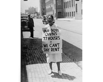 May include: A woman in a white dress holds a sign that reads "Our Boss Owns 77 Houses. We Can't Pay Rent." The sign is in black and white lettering on a white background. The woman is standing on a brick street in front of a building.