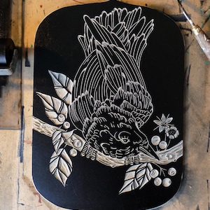 May include: A black wooden plaque with a white carved illustration of a bird perched on a branch with leaves and berries. The bird is facing downward, with its beak near the berries. The plaque has a decorative, shield-like shape.
