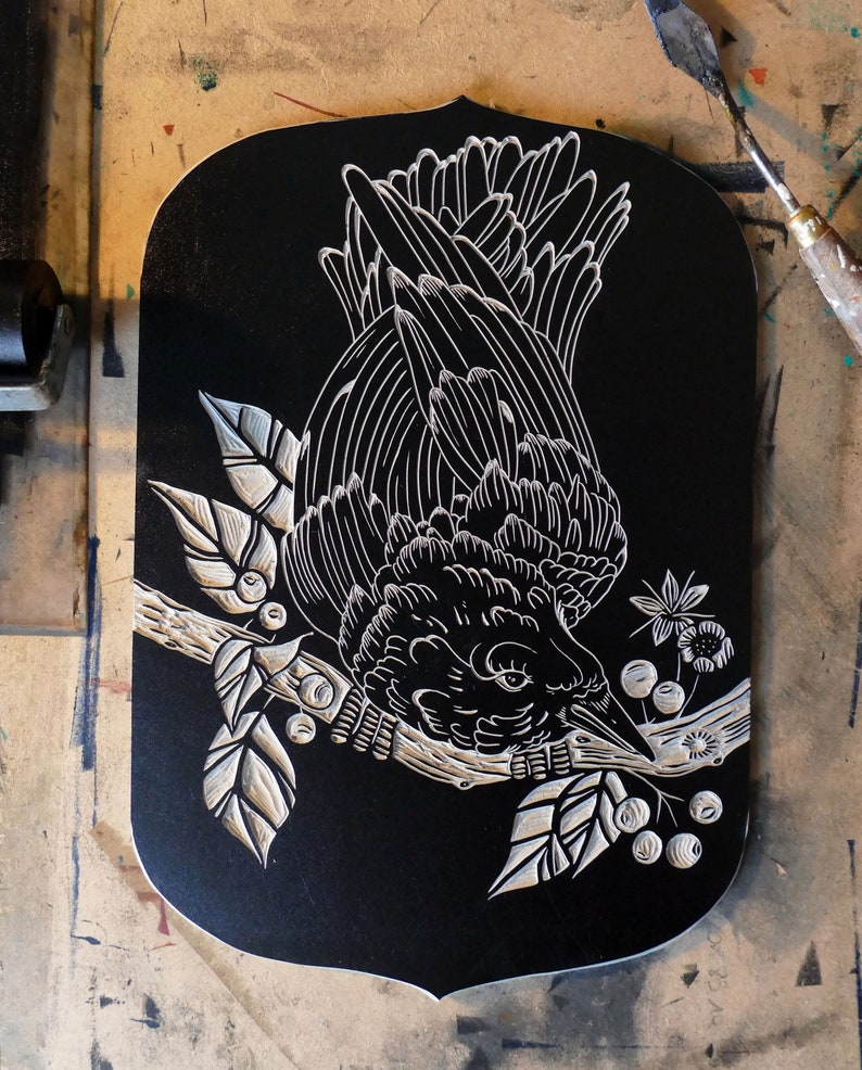May include: A black wooden plaque with a white carved illustration of a bird perched on a branch with leaves and berries. The bird is facing downward, with its beak near the berries. The plaque has a decorative, shield-like shape.