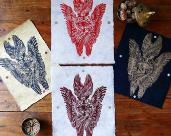 May include: Three prints of a winged eye design. The prints are on different colored paper: red, black, and cream. The eye is surrounded by a crown and wings.