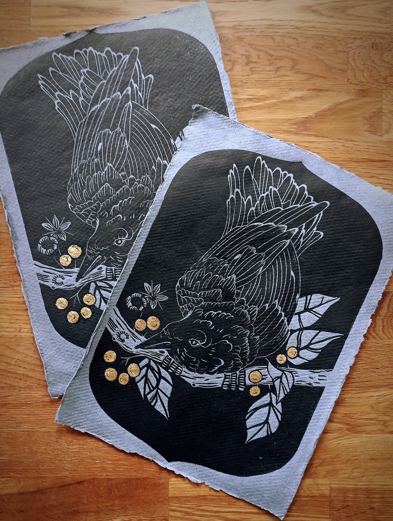 May include: Two linocut prints featuring a black bird perched on a branch with leaves and gold berries. The bird is depicted with white linework against a dark background. The prints have a textured, handmade paper appearance.