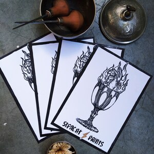 May include: Four white cards with black line drawings of a flaming chalice. The cards are stacked on top of each other. The bottom card has the text "Sleekat Prints" in black.