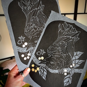 May include: Two black and gray art prints featuring a detailed illustration of a crow perched on a branch with berries. The crow is rendered in white lines against a black background. The prints have a gray border.