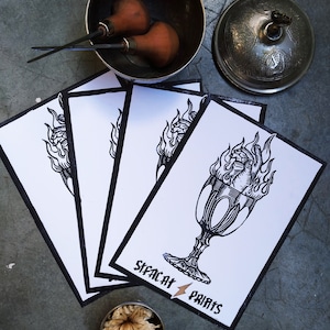 May include: A collection of art prints with a black and white illustration of a flaming chalice, with the text "SIFACAT PRINTS". The prints are surrounded by art tools and a decorative bowl.