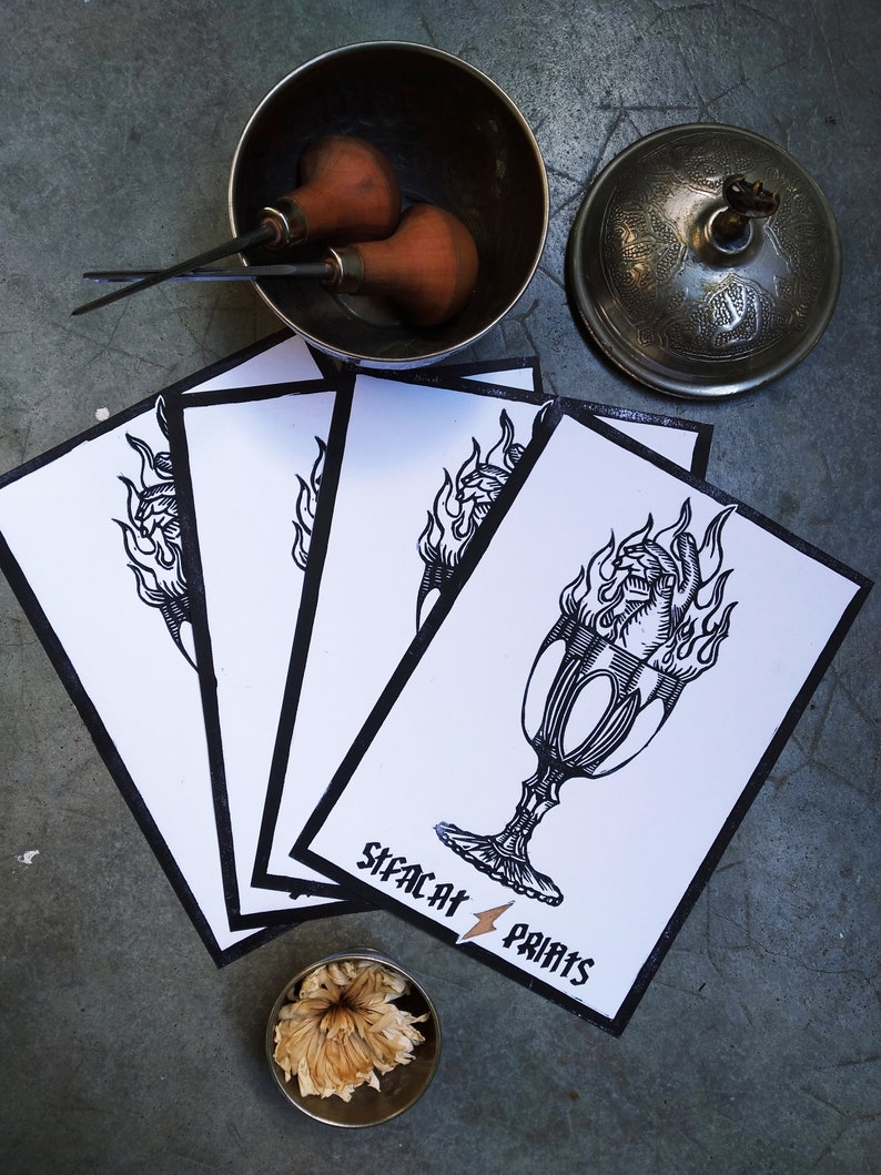 May include: A collection of art prints with a black and white illustration of a flaming chalice, with the text "SIFACAT PRINTS". The prints are surrounded by art tools and a decorative bowl.