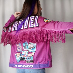 Op de afbeelding: Een roze denim jack met franjes en patches. Het jack heeft het woord "TRAVEL" in paarse letters op de rug en de woorden "THE ONLY MEMORIES" onderaan.