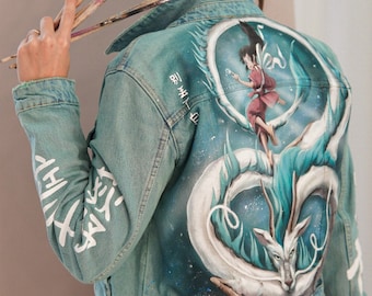 May include: A light blue denim jacket with a hand-painted design on the back. The design features a white dragon with blue accents, a character from the animated film Spirited Away, and Japanese text. The jacket has white text on the sleeves.