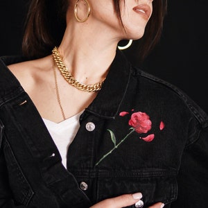 May include: A black denim jacket with a red rose painted on the front pocket. The jacket features a gold chain necklace and gold hoop earrings. The text "There" is embroidered on the sleeve.