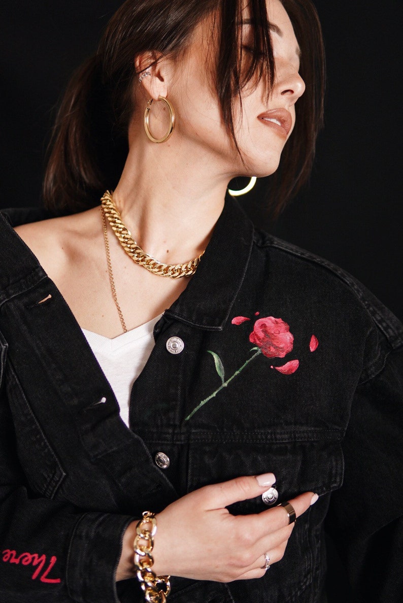 May include: A black denim jacket with a red rose painted on the front pocket. The jacket features a gold chain necklace and gold hoop earrings. The text "There" is embroidered on the sleeve.