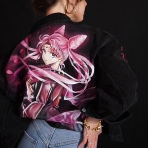 May include: A black denim jacket featuring a hand-painted image of a pink-haired anime character on the back. The character is wearing a pink dress and has a white ribbon in her hair. The jacket has a gold chain bracelet on the left wrist.