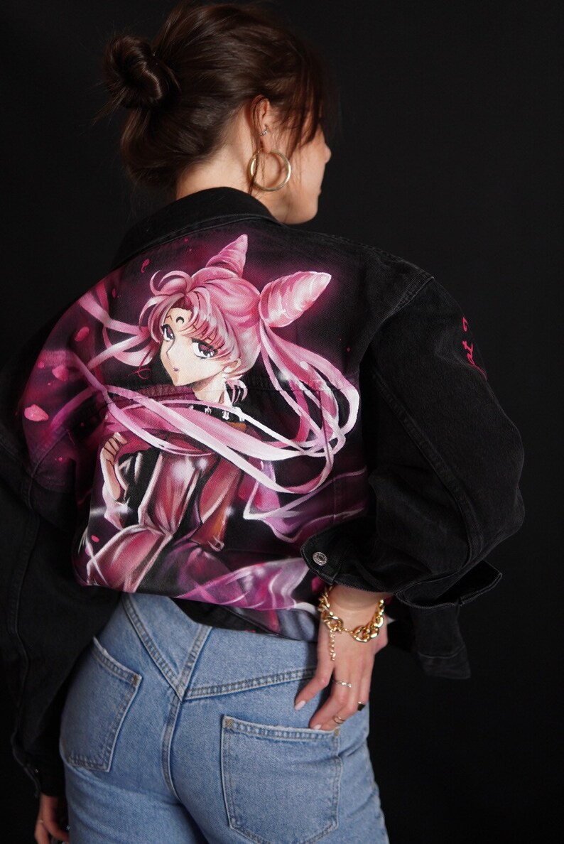 May include: A black denim jacket featuring a hand-painted image of a pink-haired anime character on the back. The character is wearing a pink dress and has a white ribbon in her hair. The jacket has a gold chain bracelet on the left wrist.