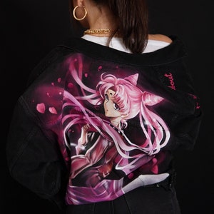 May include: Black denim jacket featuring a hand-painted image of a pink-haired anime character with a pink ribbon and a crown. The text "Sailor" is written on the jacket.