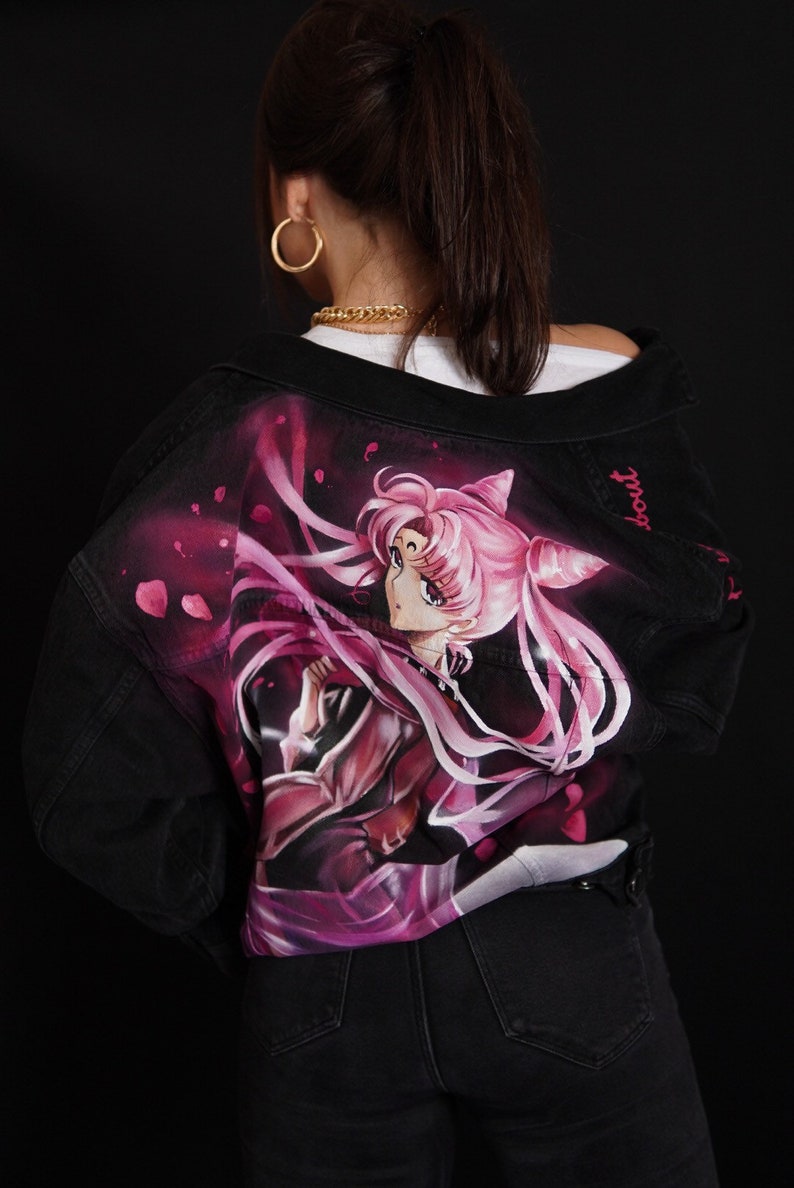 May include: Black denim jacket featuring a hand-painted image of a pink-haired anime character with a pink ribbon and a crown. The text "Sailor" is written on the jacket.