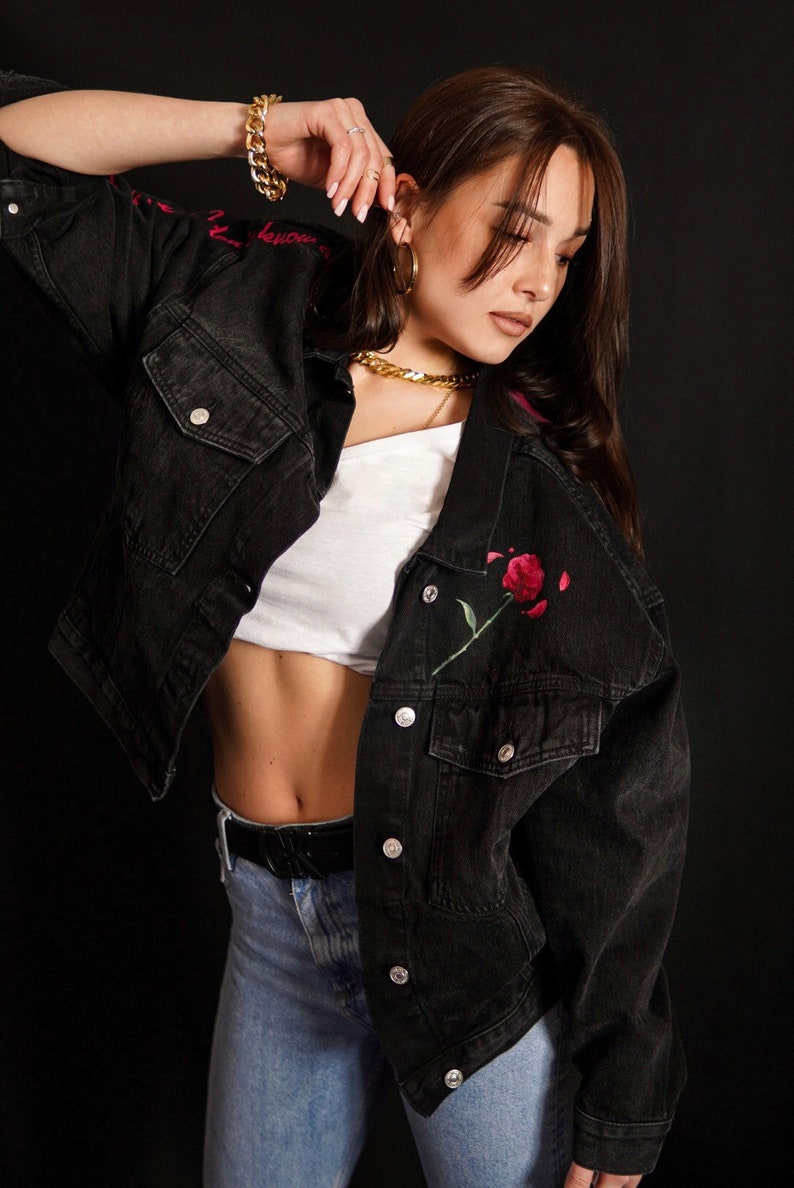 May include: A woman wearing a black denim jacket with a red rose embroidered on the front. She is also wearing a white t-shirt, blue jeans, and gold jewellery.