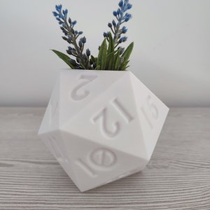 Puede incluir: Maceta de cerámica blanca con forma de dado de veinte caras con los números del 1 al 20 en relieve en los lados. La maceta está llena de una pequeña disposición de flores artificiales azules y verdes.