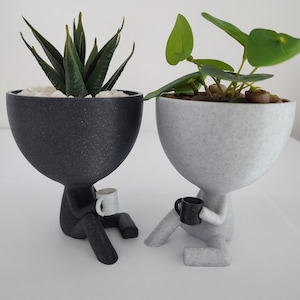 May include: Two small, 3D printed planters shaped like people, each holding a small cup. The planter on the left is black and has a succulent plant inside. The planter on the right is white and has a leafy plant inside.